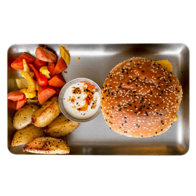 Top-down view of a sesame seed burger, roasted potatoes, mixed vegetables, and dipping sauce on a metal tray.